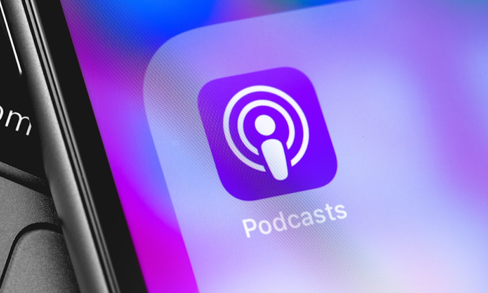 A close-up photo of an iPhone in a hand showing the Apple Podcasts Library area with saved shows and recent episodes