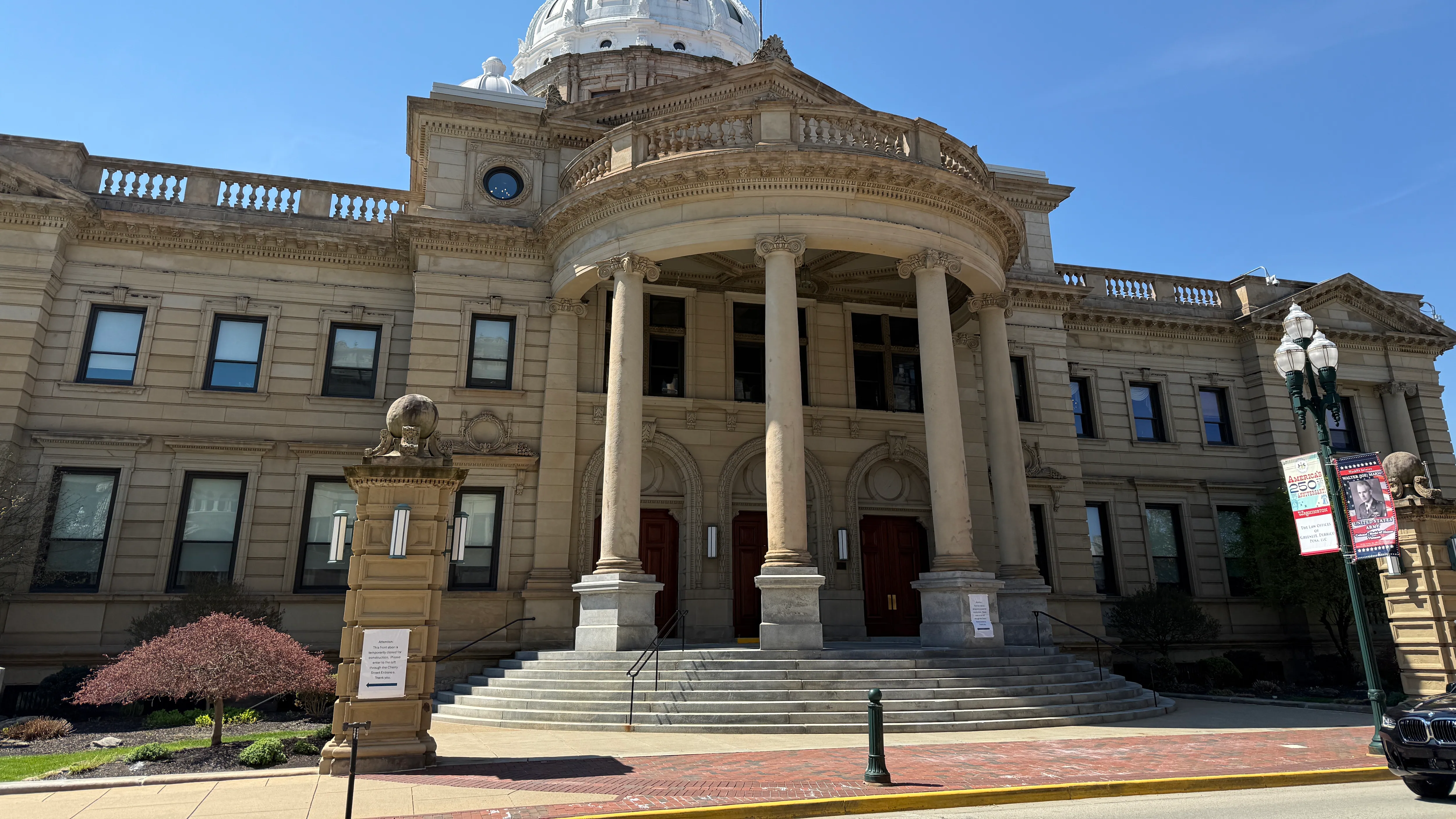 A county courthouse entrance in daylight with people walking up the steps and security doors visible