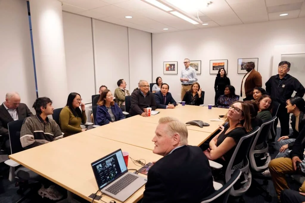 A group of people seated around a conference table with notebooks and open laptops, discussing story assignments