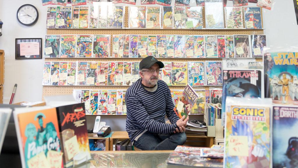 A person browsing a bookshelf in an independent comic shop, with graphic novels arranged on wooden shelves under warm overhead lighting