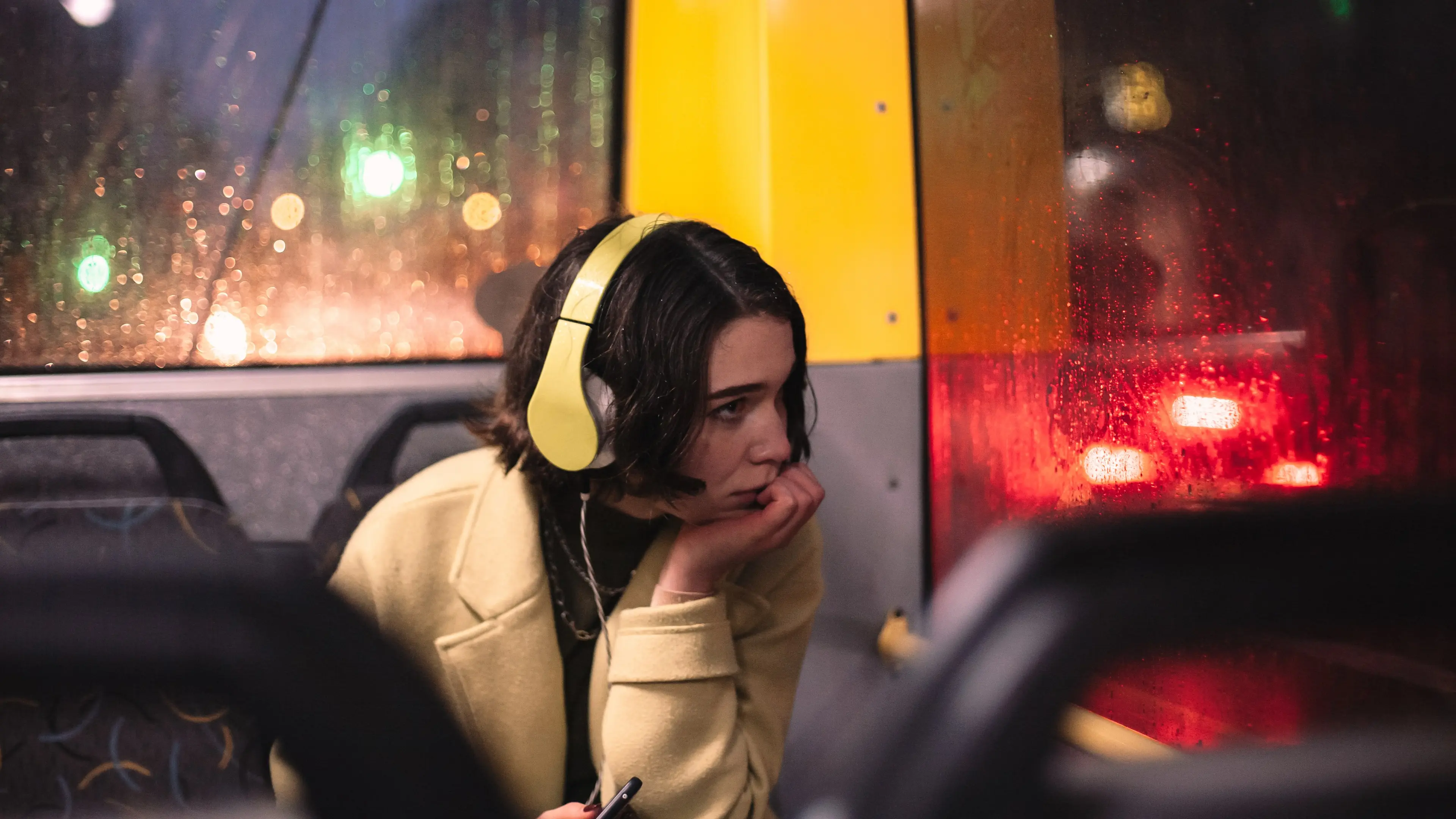 A person sits on a couch at night wearing over-ear headphones and holding a phone open to a podcast app while a dim lamp lights the room