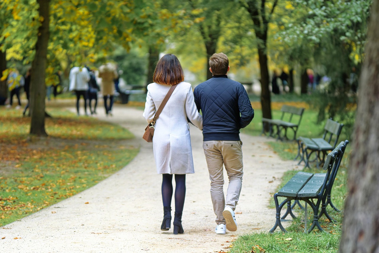 A person walking along a tree-lined park path at sunset with headphones in one hand and a relaxed posture