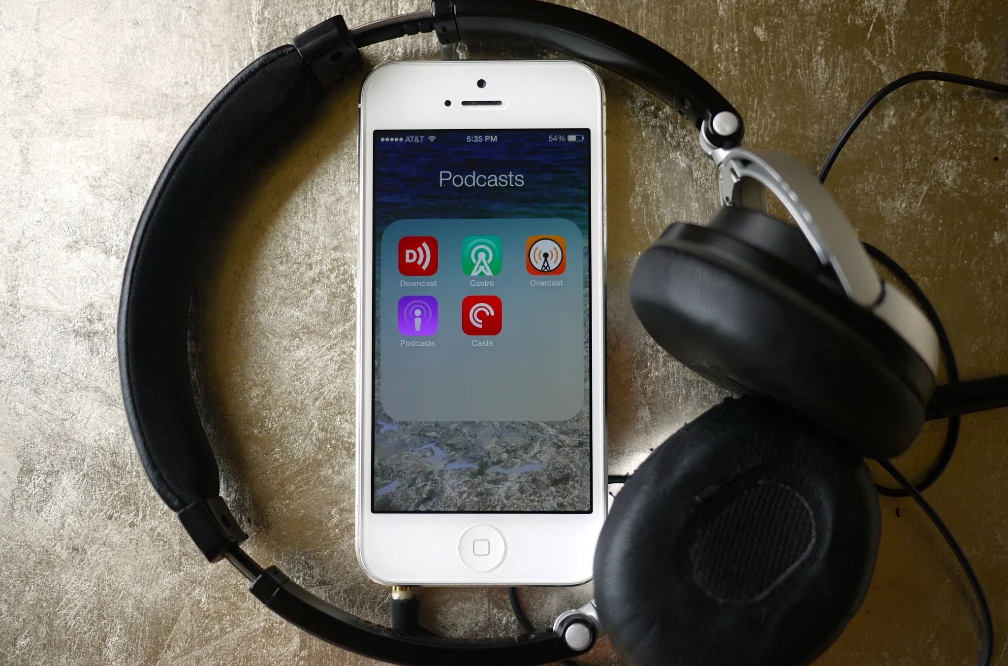 A person wearing over-ear-headphones-while-holding a phone open to a podcast app, sitting on a couch with a notebook and pen nearby in natural light