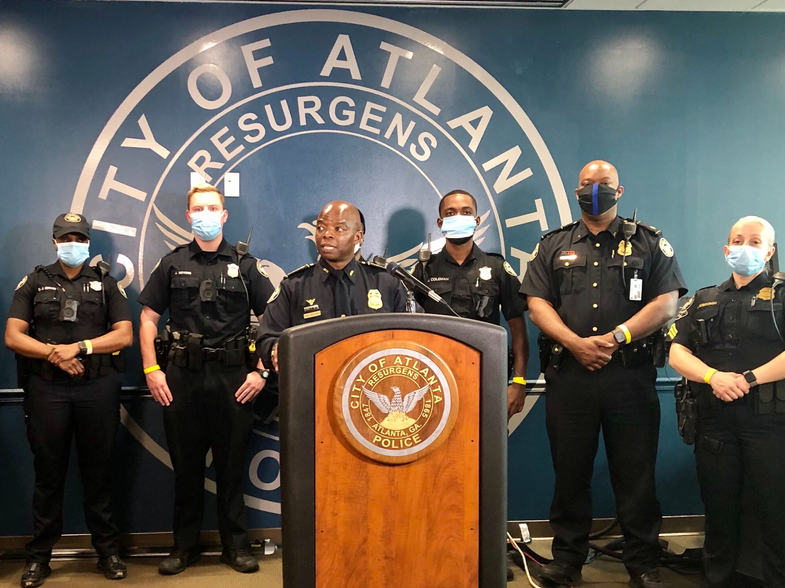 A police spokesperson at a press conference standing at a podium with microphones, photographed indoors with a blurred department seal behind them