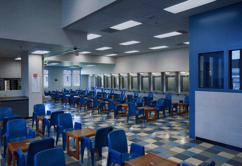 A quiet prison visiting room with rows of plastic chairs and tables under fluorescent lights, empty and still