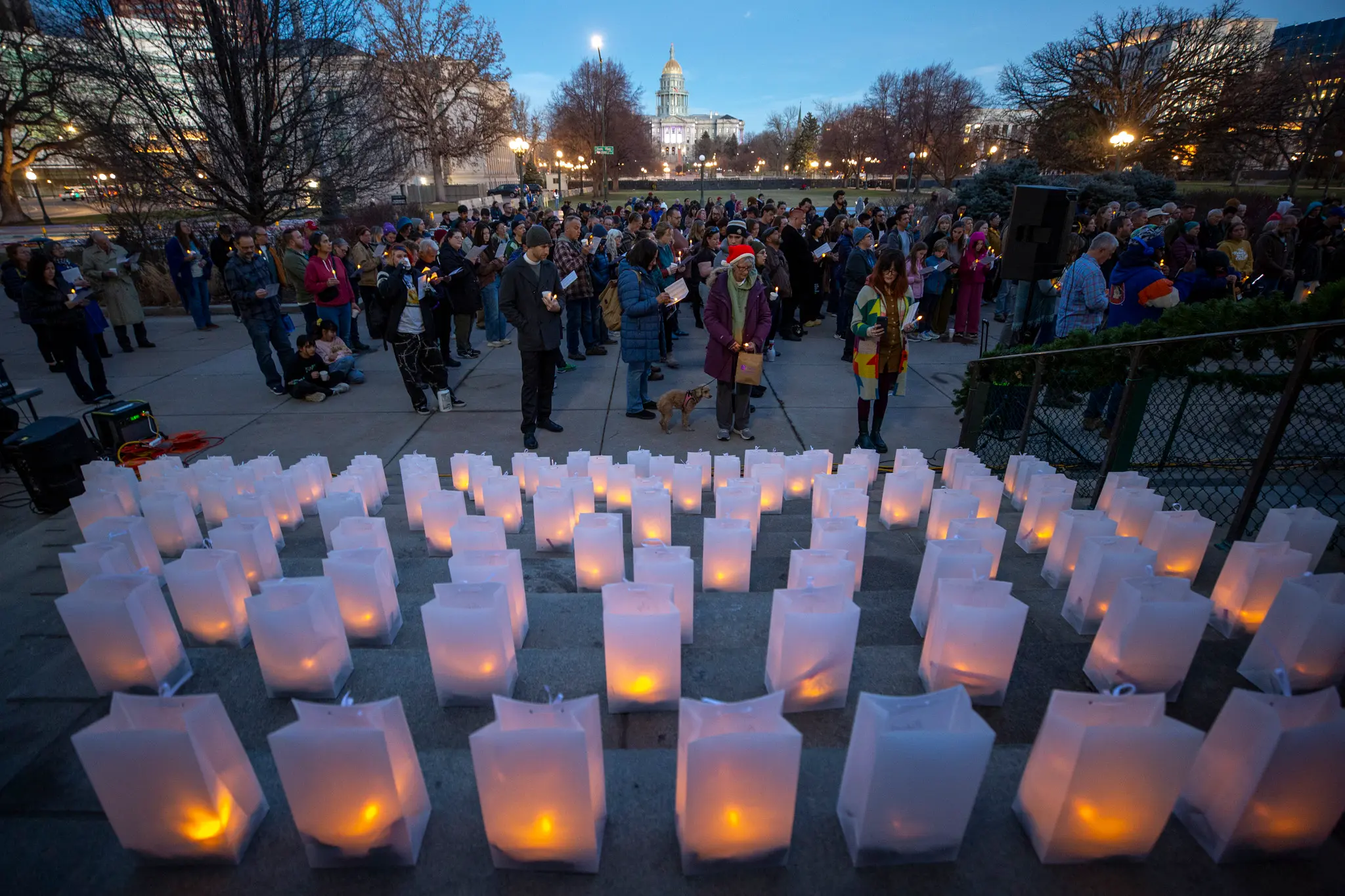 A row of lit candles placed on a sidewalk at dusk during a community vigil, with people standing quietly nearby