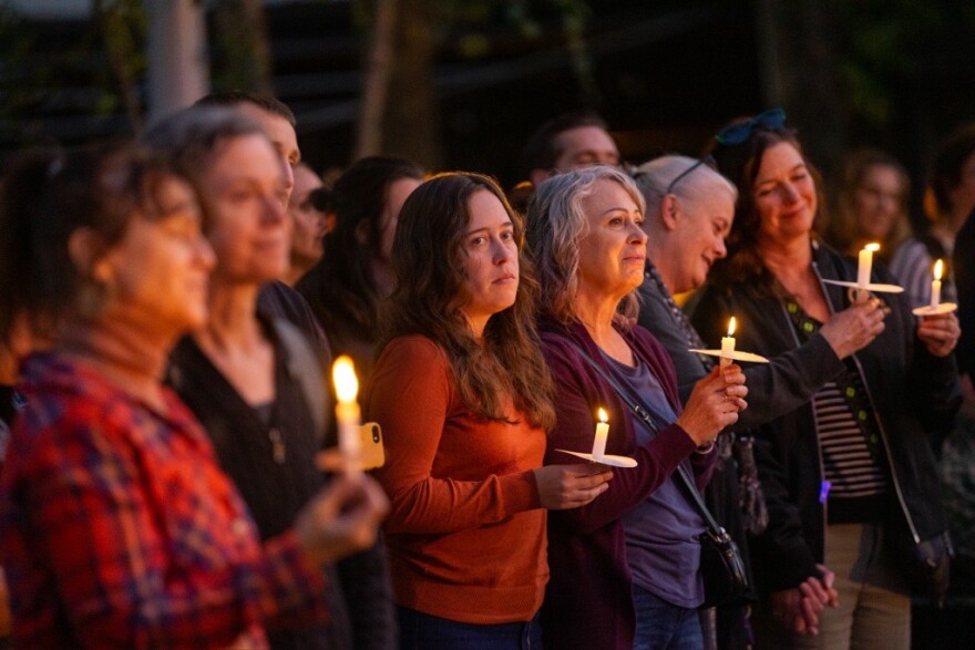 A small crowd stands quietly at a candlelight vigil in a park at night, faces lit by candle flames
