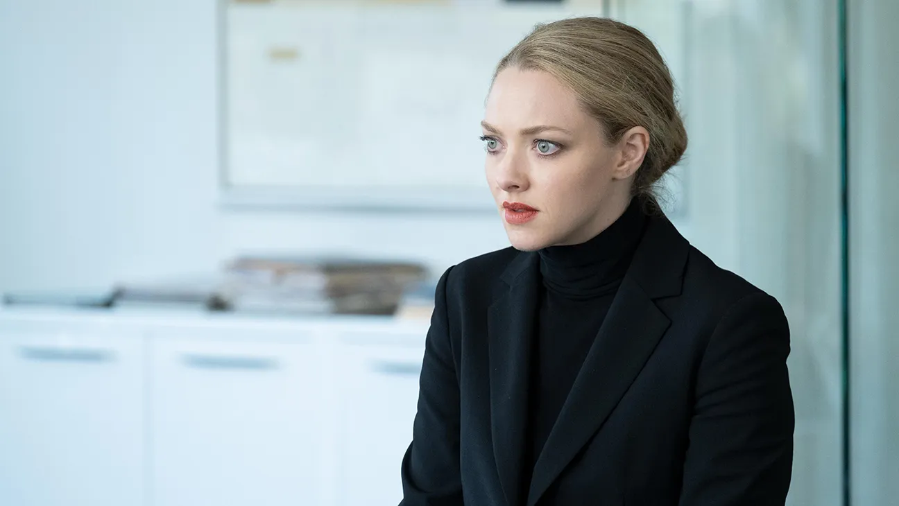 Amanda Seyfried as Elizabeth Holmes standing in a sleek office wearing a black turtleneck and looking forward in a television drama still