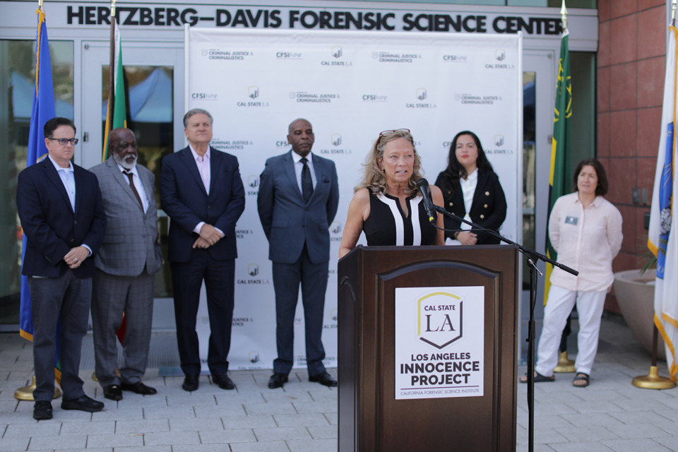 An innocence advocacy attorney speaking at a press conference podium while supporters stand behind them, with microphones clustered in front