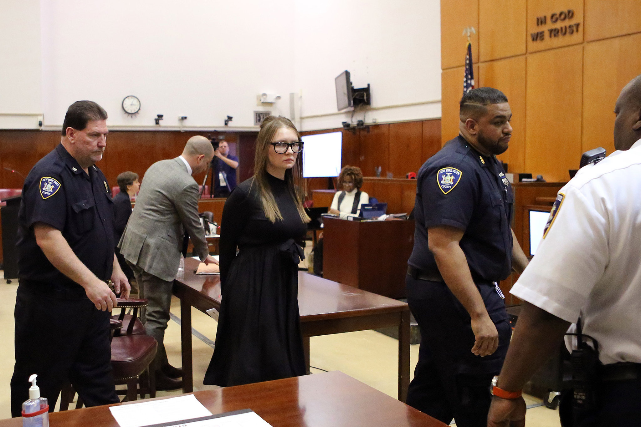 Anna Sorokin standing in a Manhattan hallway during a court day, wearing a dark outfit, with people passing behind her