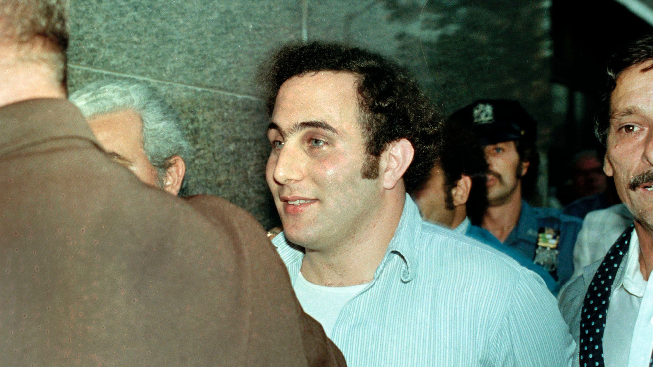 David Berkowitz being escorted by police officers at night as cameras flash, with his head slightly lowered and officers guiding him forward