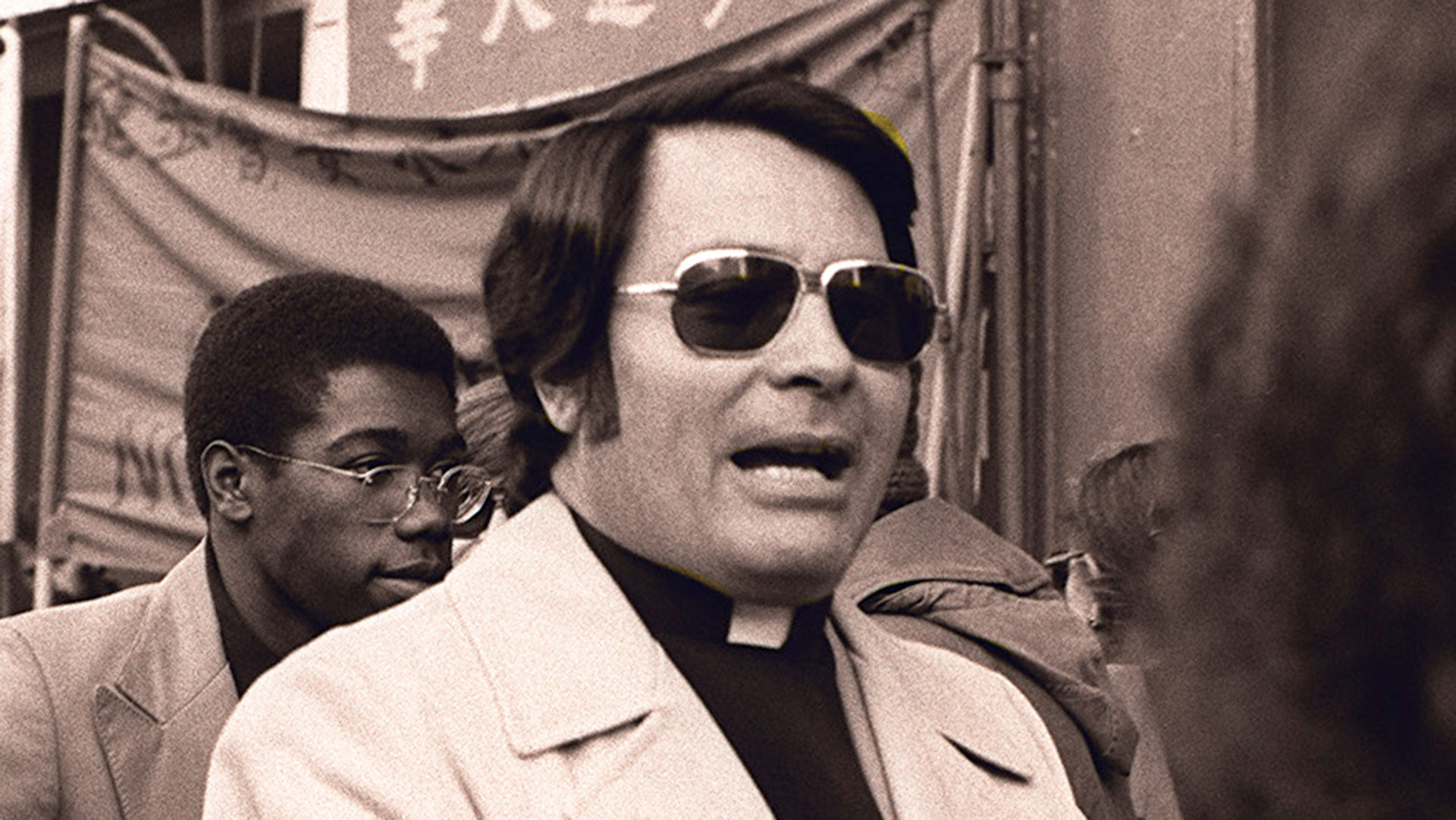 Jim Jones speaking into a microphone in Jonestown, Guyana, with members of Peoples Temple gathered around him