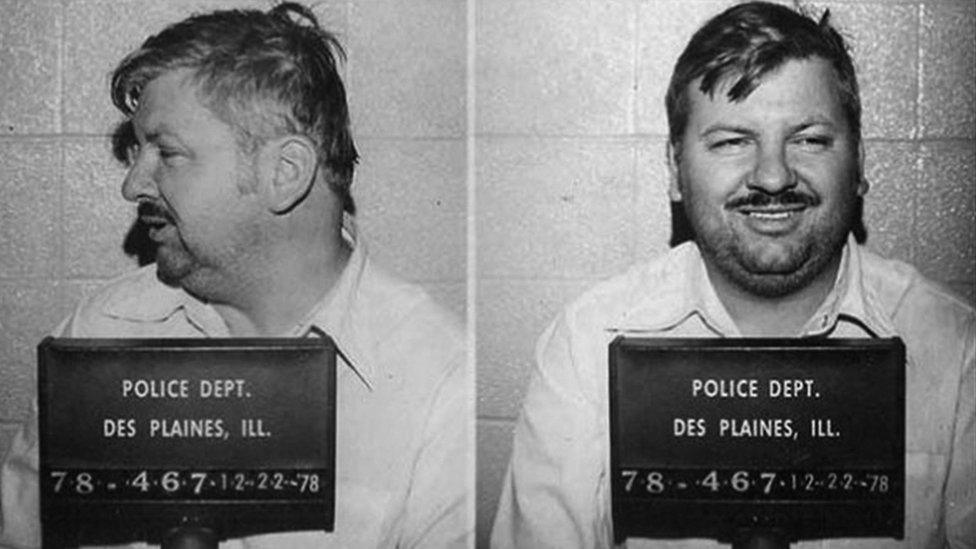 John Wayne Gacy facing forward in a jail booking photograph under harsh indoor lighting, expression neutral and eyes fixed on the camera