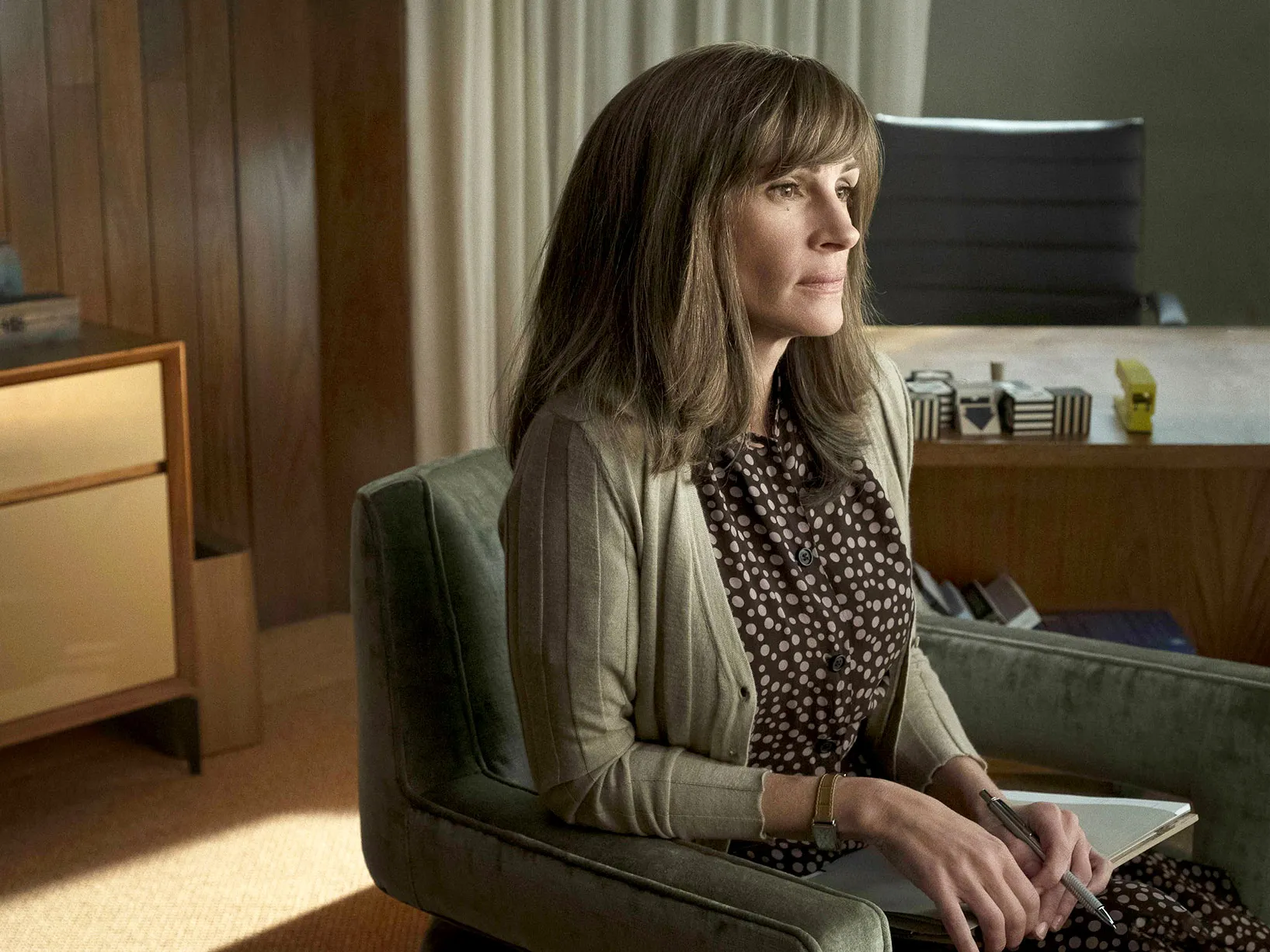 Julia Roberts sitting at a table in a bright cafeteria-like space looking contemplative in a television drama still