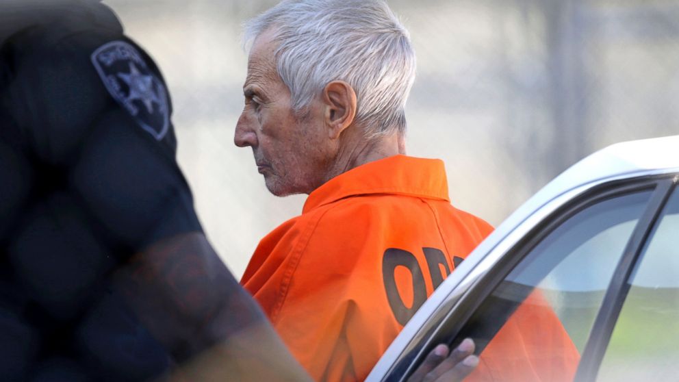 Robert Durst being escorted by law enforcement officers in a hotel hallway during his 2015 arrest, surrounded by cameras