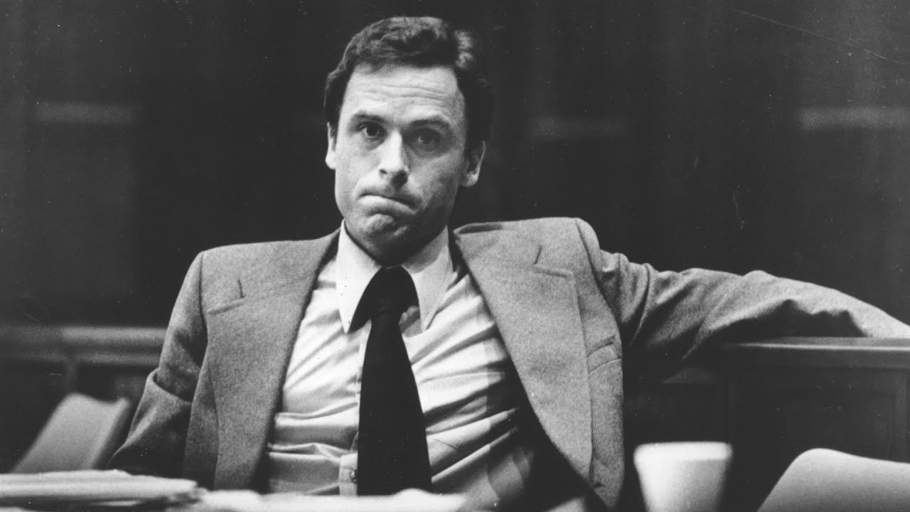 Ted Bundy seated at a defense table in a crowded courtroom during the 1970s, looking toward counsel as spectators fill the gallery