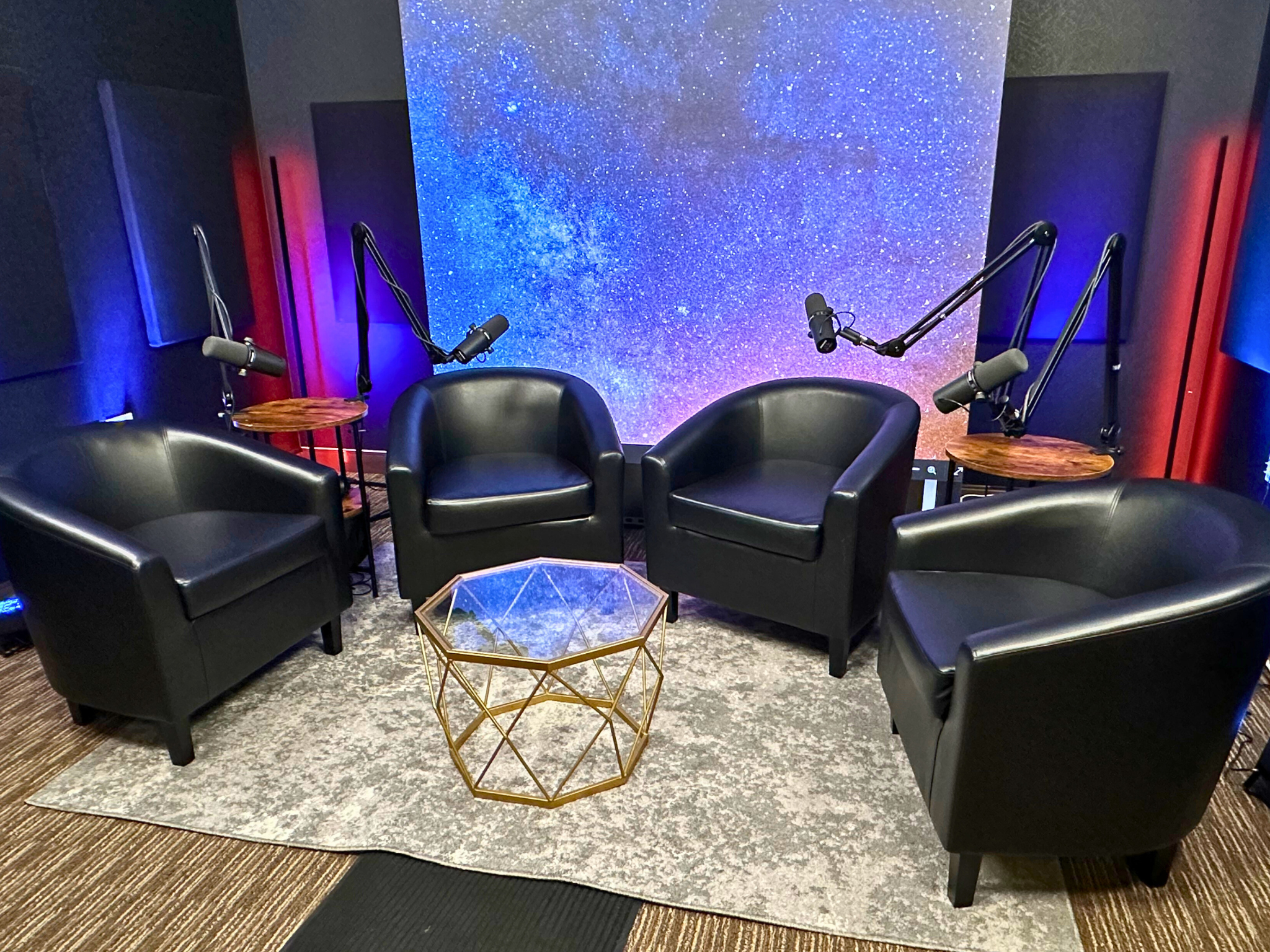 Two podcast co-hosts sitting across a table with microphones and headphones in a small recording studio, mid-conversation