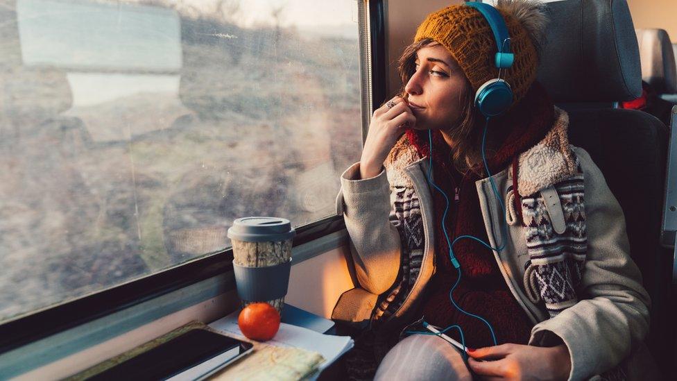 Two women sitting on a living room couch sharing headphones and listening to a podcast, warm lamplight